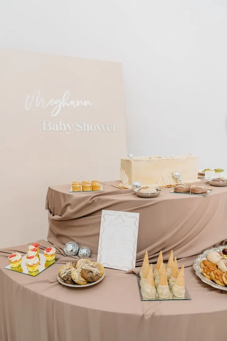 Buffet de baby shower sur drapés taupe devant un panneau rose poudré au prénom de la future maman